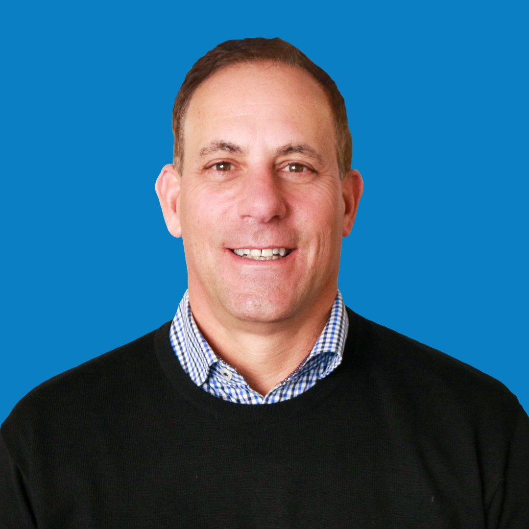Portrait of a smiling man with short dark hair, wearing a black sweater over a blue and white checkered button-down shirt, against a medium blue circular background.