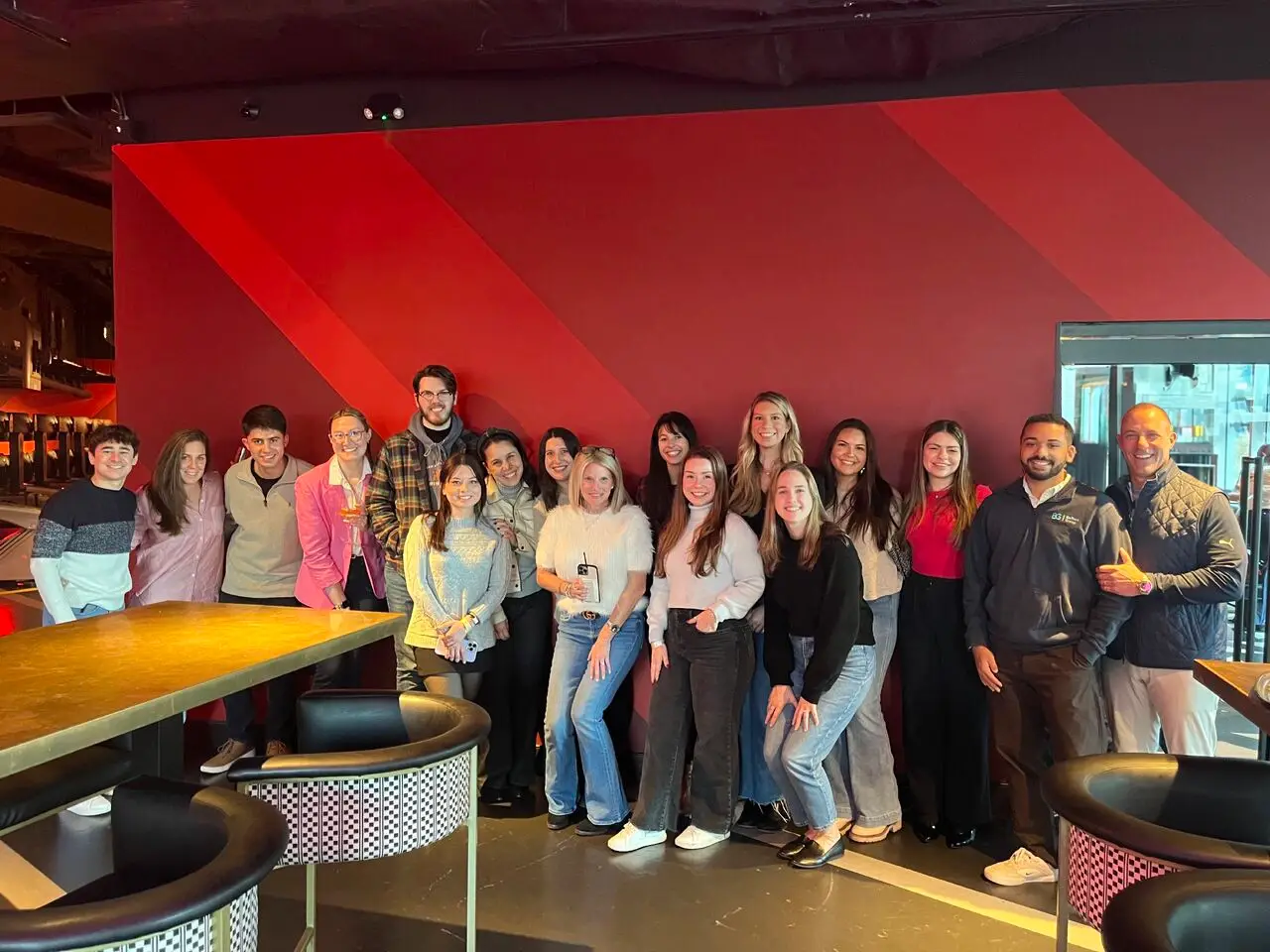 A group of fifteen smiling colleagues posing for a photo against a deep red, diagonal-striped wall in a modern lounge or restaurant setting. The group is dressed in a mix of business casual and smart attire. In the foreground, there are high-top tables with checkered-patterned barrel chairs.